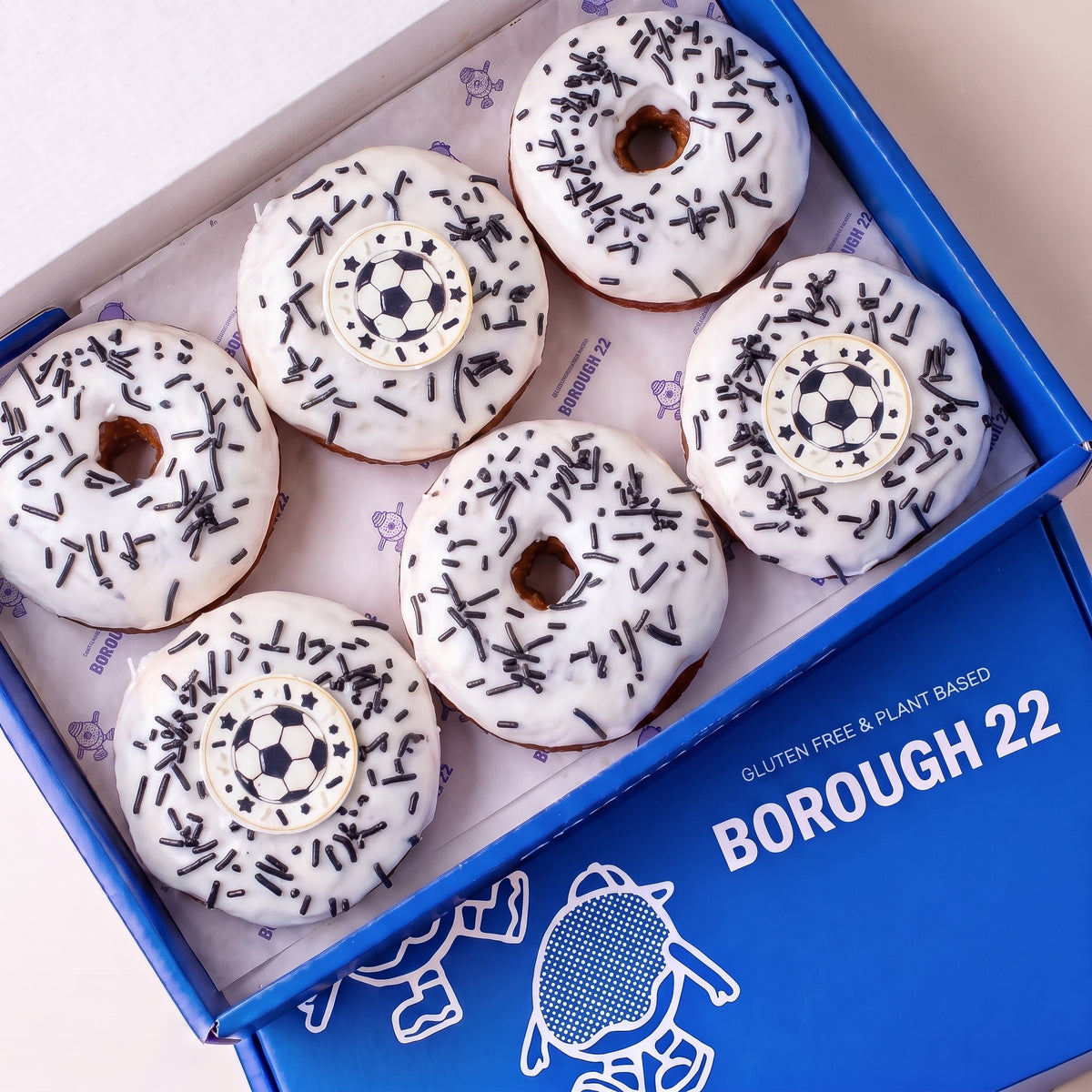 Gluten free vegan football doughnuts with white icing, black strand sprinkles and football toppers in a Borough 22 box