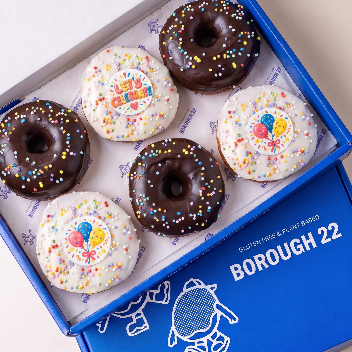 Gluten free vegan party doughnuts with chocolate and white icing, rainbow sprinkles and balloon toppers in a Borough 22 box